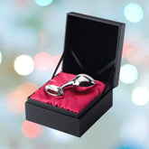 The njoy Pure Butt Plug Large by Njoy, made of stainless steel and polished to a mirror finish, is elegantly displayed on a red satin cushion inside an open black gift box. The blurred background, adorned with multicolored circular lights, adds a festive touch to the scene.